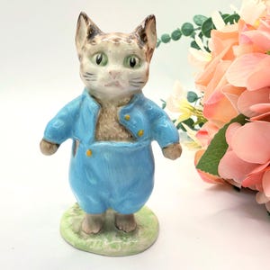 May include: A ceramic figurine of a cat wearing a blue outfit with gold buttons. The cat has green eyes and a light brown and white face. The figurine is standing on a green base, with a bouquet of pink flowers in the background.