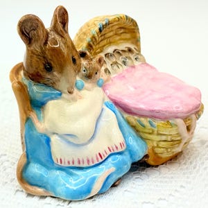 May include: A ceramic figurine of a mother mouse and two babies. The mother mouse is brown and wears a blue dress and white shawl. The babies are in her arms. A pink-covered basket is behind them. The figurine is on a white surface.