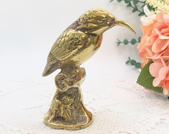 May include: A brass bird figurine perched on a decorative base. The bird has detailed feather and wing patterns, with a long, curved beak. The base features leaf and floral designs. The figurine is a golden color.