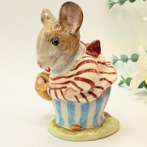 May include: A ceramic mouse figurine wearing a striped shirt and blue striped trousers, holding a basket. The mouse has brown fur, a red bow, and stands on a green base. A decorative item.