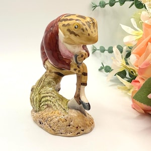 May include: A ceramic figurine of a lizard-like creature, perched on a textured base. The figure has a tan and brown spotted head and wears a burgundy coat. The base is a mix of tan, green, and brown. Decorative item.