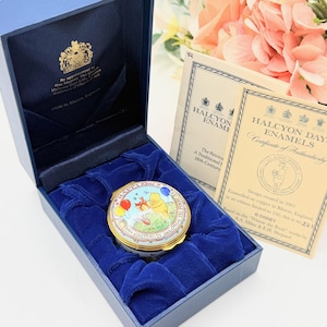May include: A blue box with a velvet interior holds a round, gold-rimmed enamel trinket box. The box features a colorful Winnie the Pooh design. Two cream-colored cards with gold lettering and a floral arrangement are in the background.
