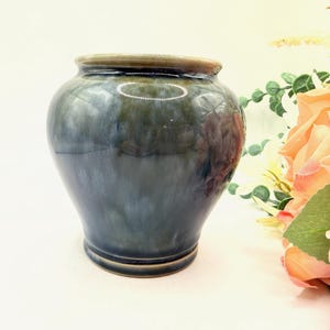 May include: A small, rounded ceramic vase featuring a glossy, multi-toned glaze that transitions from deep navy blue to muted sage green. The vessel has a wide, curved body, a short neck, and a smooth, circular rim with a subtle brown edge finish.