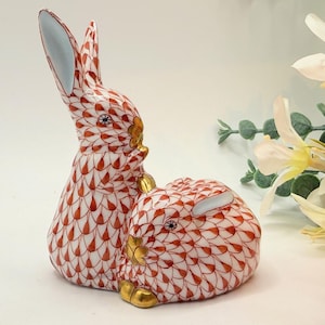 May include: Two ceramic rabbit figurines. The rabbits are white with a red and gold pattern. One rabbit is upright, and the other is lying down. The ears of the upright rabbit have a light blue interior. The figurines have gold accents.