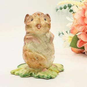May include: A ceramic figurine of a hamster, standing upright on a green and yellow leaf base. The hamster is painted in shades of brown, tan, and pink. The figurine has detailed features, including small ears and whiskers. Decorative item.