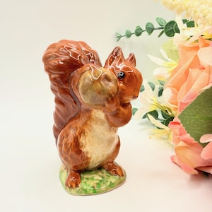 May include: A ceramic squirrel figurine in shades of brown, tan, and cream, holding a nut. The squirrel has a bushy tail and is posed on a patch of green grass. The figurine is a decorative item.