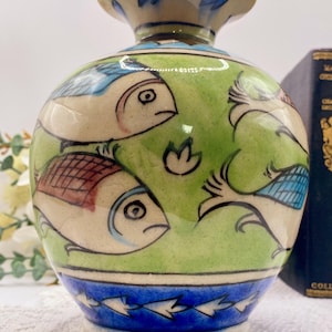 May include: A hand-painted ceramic vase with a bulbous body and a narrow neck. The vase features a green background with fish illustrations in shades of blue, red, and white. The base and neck are adorned with blue and white accents.