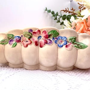 May include: Cream-coloured ceramic planter with a scalloped design. The planter features colourful, raised floral accents in shades of pink, blue, and purple, with green leaves. A decorative piece for home decor.