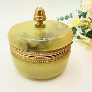 May include: A round, pale green stone trinket box with a polished finish and natural veining. The lid features a central brass acorn-shaped finial, and the base is encircled by a decorative, textured brass band that connects the lid to the container.