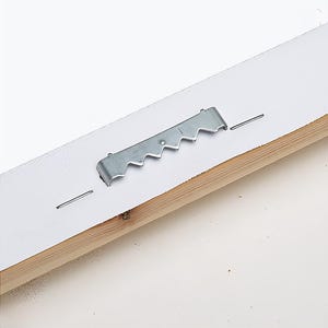 Pu&ograve; includere: Primo piano del retro di un'opera d'arte incorniciata. Un gancio a dente di sega in metallo &egrave; attaccato al supporto bianco con piccoli chiodi. La cornice in legno &egrave; visibile in basso. Lo sfondo &egrave; bianco.