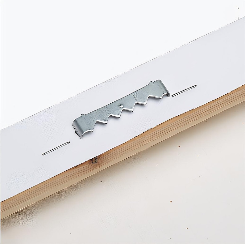 Pu&ograve; includere: Primo piano del retro di un'opera d'arte incorniciata. Un gancio a dente di sega in metallo &egrave; attaccato al supporto bianco con piccoli chiodi. La cornice in legno &egrave; visibile in basso. Lo sfondo &egrave; bianco.