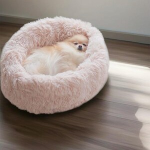May include: A fluffy pink dog bed with a small dog sleeping inside. The bed is made of soft, plush material and has a round shape.