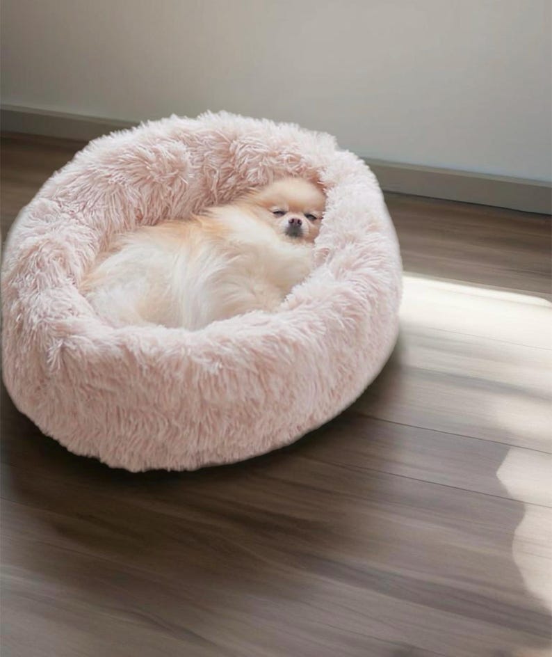 May include: A fluffy pink dog bed with a small dog sleeping inside. The bed is made of soft, plush material and has a round shape.