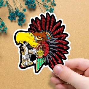 Puede incluir: Una colorida pegatina con un tocado de águila azteca estilizado sobre una calavera. El diseño incluye acentos rojos, amarillos, negros y verdes. La pegatina se sostiene sobre un fondo marrón claro. El texto en la imagen dice "DIFERENTES TAMAÑOS DISPONIBLES."