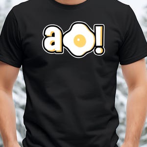 May include: Black t-shirt featuring a graphic of a fried egg with the letters "a o!" in a playful font. The egg design has a yellow yolk and a white outline, with a black border.
