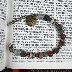 May include: A beaded bracelet with a bronze-colored charm and clasp. The bracelet features a variety of beads in shades of gray, brown, and red, including lava rock and patterned stones. The charm has the text "GEN 28:15".