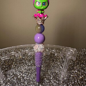May include: Purple and green pen with a glittery purple body and a crown topped with a green and pink character figurine.
