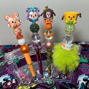 May include: Four decorative pens with character-themed toppers. The pens feature orange, purple, and green barrels, adorned with beads and character figures. The toppers include a monkey, Minnie Mouse, a chocolate bar character, and a flower character. The pens are displayed in a clear plastic holder.