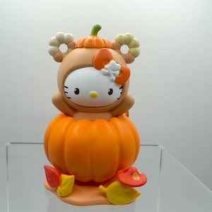 May include: A Hello Kitty figurine dressed in a tan bear costume with a pumpkin hat, sitting inside a large orange pumpkin. The character features a white face, yellow nose, and an orange bow with a white skull icon. Includes autumn leaves and a mushroom.