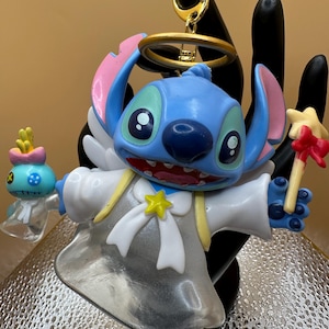 Puede incluir: Figura azul de Stitch con alas de &aacute;ngel, un halo y un acento en forma de estrella. La figura sostiene una varita en forma de estrella y una peque&ntilde;a figura alien&iacute;gena azul. Un llavero dorado est&aacute; adjunto en la parte superior.