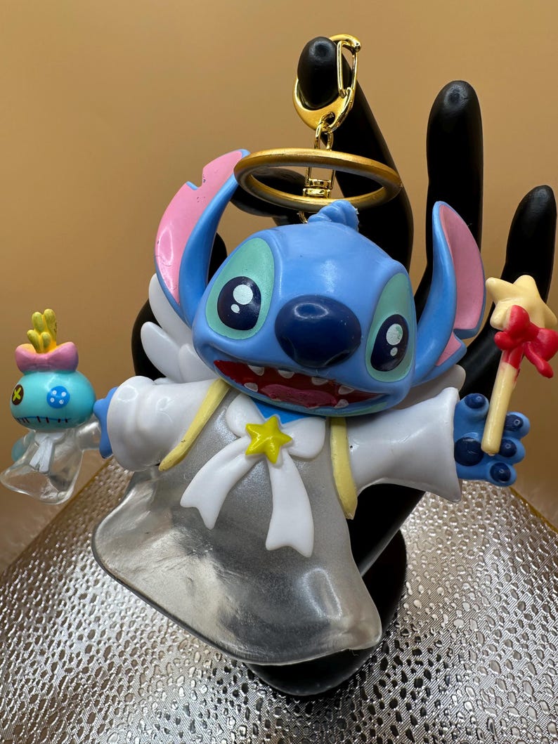 Puede incluir: Figura azul de Stitch con alas de &aacute;ngel, un halo y un acento en forma de estrella. La figura sostiene una varita en forma de estrella y una peque&ntilde;a figura alien&iacute;gena azul. Un llavero dorado est&aacute; adjunto en la parte superior.