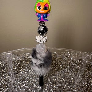 May include: A black and white pen with a fluffy white and black pom pom on top. The pen is topped with a black and white beaded charm and a small plastic figurine with green and orange hair, blue clothing, and a white and black face.