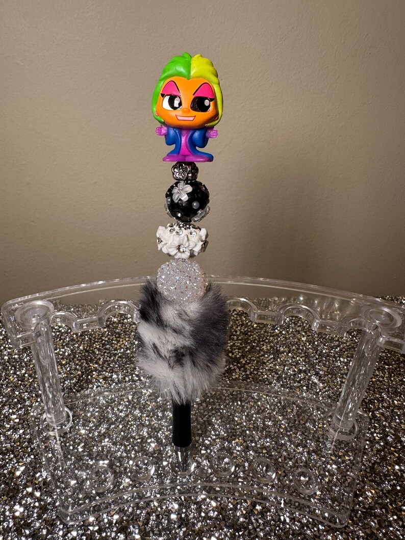 May include: A black and white pen with a fluffy white and black pom pom on top. The pen is topped with a black and white beaded charm and a small plastic figurine with green and orange hair, blue clothing, and a white and black face.