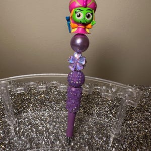 May include: Purple and green pen with a sparkly purple design and a green character with a pink turban on top. The pen is in a clear plastic pen holder.