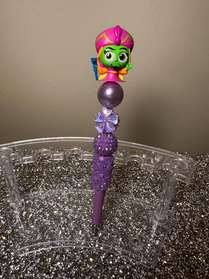 May include: Purple and green pen with a sparkly purple design and a green character with a pink turban on top. The pen is in a clear plastic pen holder.