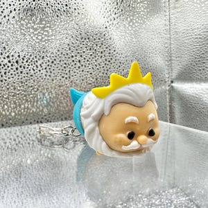 May include: A small, rounded figurine keychain depicting a character with a tan face, white hair, a white mustache, and a yellow crown. The character has a blue tail section and a silver metal keychain ring attached to the back of the figure.