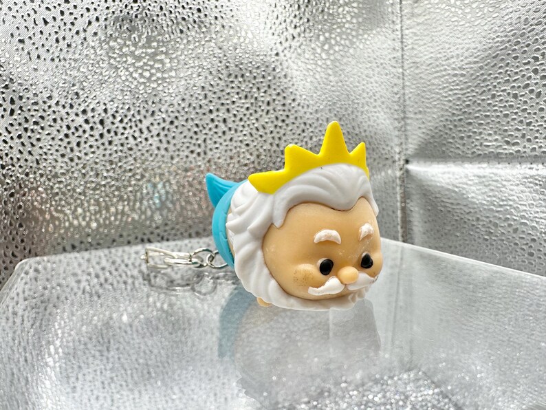 May include: A small, rounded figurine keychain depicting a character with a tan face, white hair, a white mustache, and a yellow crown. The character has a blue tail section and a silver metal keychain ring attached to the back of the figure.