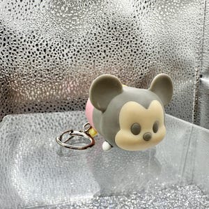 May include: A gray and cream-colored silicone keychain shaped like a stylized cartoon mouse head with large rounded ears, a small nose, and oval eyes. The character figure is attached to a circular silver-toned metal split ring for keys.