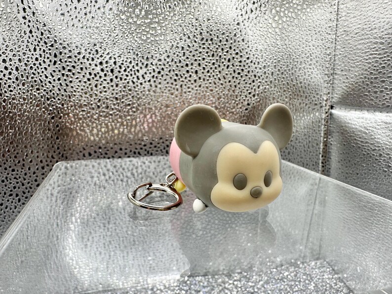 May include: A gray and cream-colored silicone keychain shaped like a stylized cartoon mouse head with large rounded ears, a small nose, and oval eyes. The character figure is attached to a circular silver-toned metal split ring for keys.