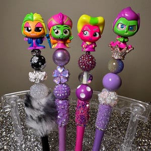 May include: Four purple and pink pens with colorful cartoon characters on top. The pens are decorated with beads, glitter, and fur.