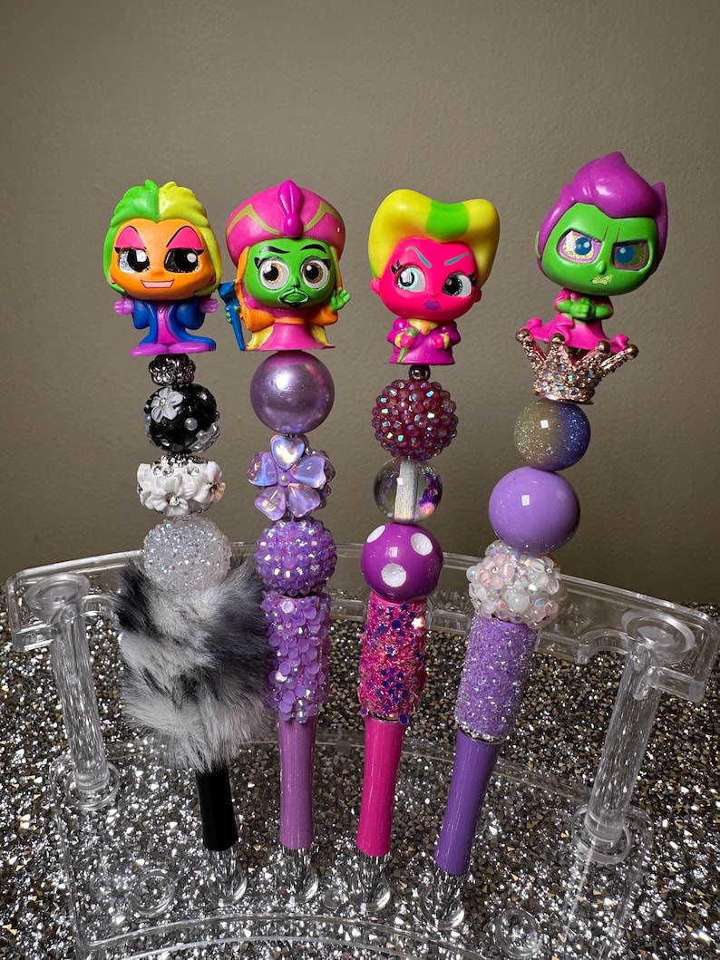 May include: Four purple and pink pens with colorful cartoon characters on top. The pens are decorated with beads, glitter, and fur.