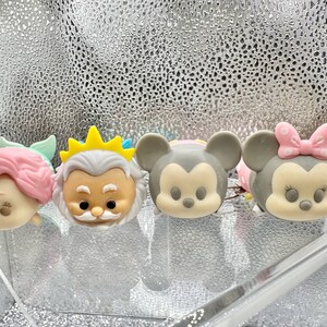 May include: Four small, rounded character figurines arranged in a row. The first is a light-skinned figure with pink hair, the second is an older figure with a white beard and yellow crown, the third is a gray mouse-like figure, and the fourth is a gray mouse-like figure with a pink polka-dot bow.