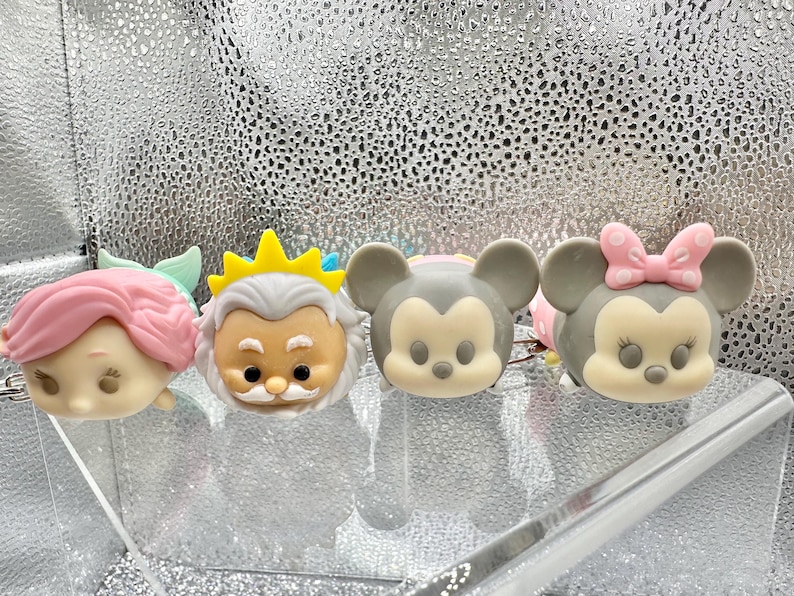 May include: Four small, rounded character figurines arranged in a row. The first is a light-skinned figure with pink hair, the second is an older figure with a white beard and yellow crown, the third is a gray mouse-like figure, and the fourth is a gray mouse-like figure with a pink polka-dot bow.