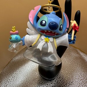 Puede incluir: Una figura azul de Stitch con orejas rosas, un halo y alas de &aacute;ngel. La figura sostiene una criatura azul y amarilla en una mano y una varita con forma de estrella en la otra. Lleva una t&uacute;nica blanca con una estrella y un lazo amarillos.