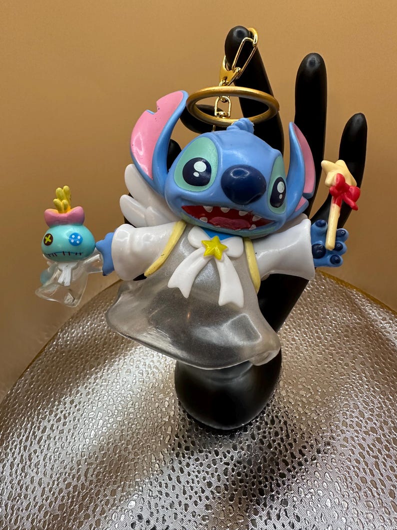 Puede incluir: Una figura azul de Stitch con orejas rosas, un halo y alas de &aacute;ngel. La figura sostiene una criatura azul y amarilla en una mano y una varita con forma de estrella en la otra. Lleva una t&uacute;nica blanca con una estrella y un lazo amarillos.