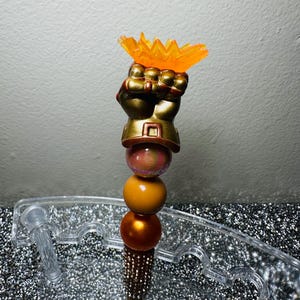 May include: A decorative pen featuring a gold-colored fist holding an orange flame-shaped accent. The pen has a series of colorful beads and a jeweled barrel. The pen is designed to be a unique writing instrument.