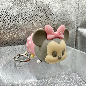 May include: A gray and cream-colored character keychain featuring a pink bow with white polka dots. The character has large round ears, a rounded face, and a small nose. A silver-toned metal split ring is attached to the side of the figure.