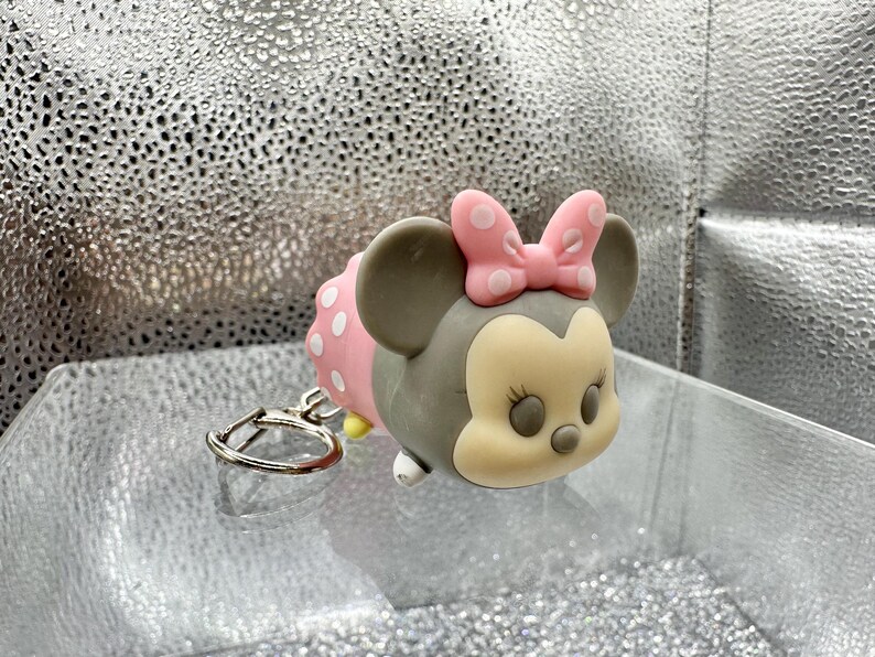 May include: A gray and cream-colored character keychain featuring a pink bow with white polka dots. The character has large round ears, a rounded face, and a small nose. A silver-toned metal split ring is attached to the side of the figure.