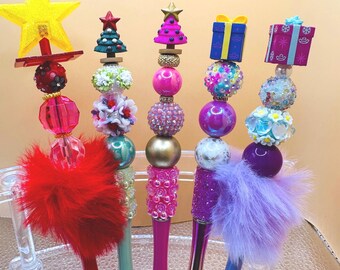 May include: A collection of decorative pens with festive Christmas-themed toppers. Each pen features a unique design, including a star, Christmas tree, and gift box, with colorful beads and feather accents. The pens are displayed in a clear holder.