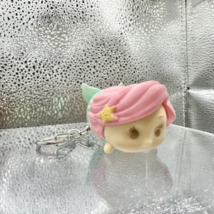 May include: A keychain featuring a stylized, rounded character head with light pink hair, a small yellow star accessory, and a mint green fin-like detail. The figure has a smooth, matte finish with simple facial features and is attached to a silver metal ring.