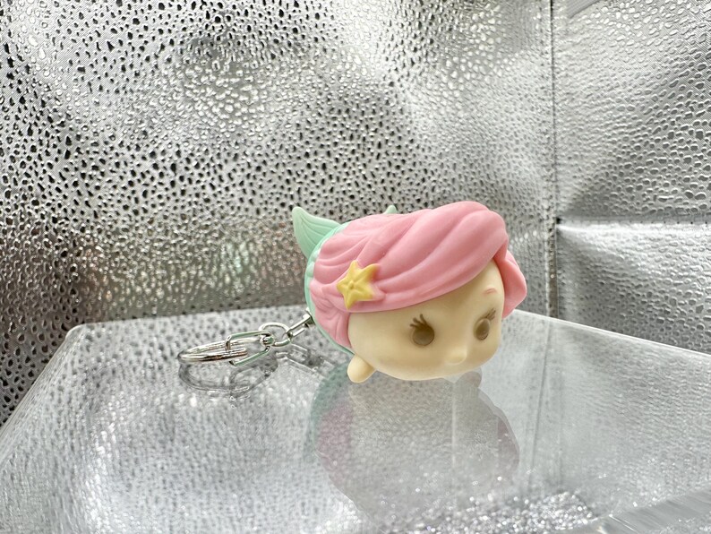May include: A keychain featuring a stylized, rounded character head with light pink hair, a small yellow star accessory, and a mint green fin-like detail. The figure has a smooth, matte finish with simple facial features and is attached to a silver metal ring.