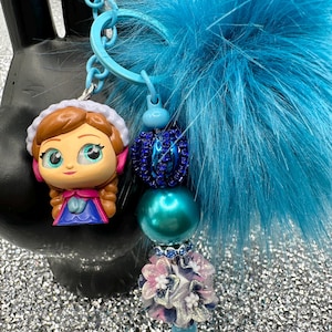 May include: A keychain featuring a cartoon character figurine with brown hair and a blue dress, a large bright blue faux fur pom-pom, and a strand of beads including a dark blue rhinestone bead, a teal pearl bead, and a floral-shaped bead.