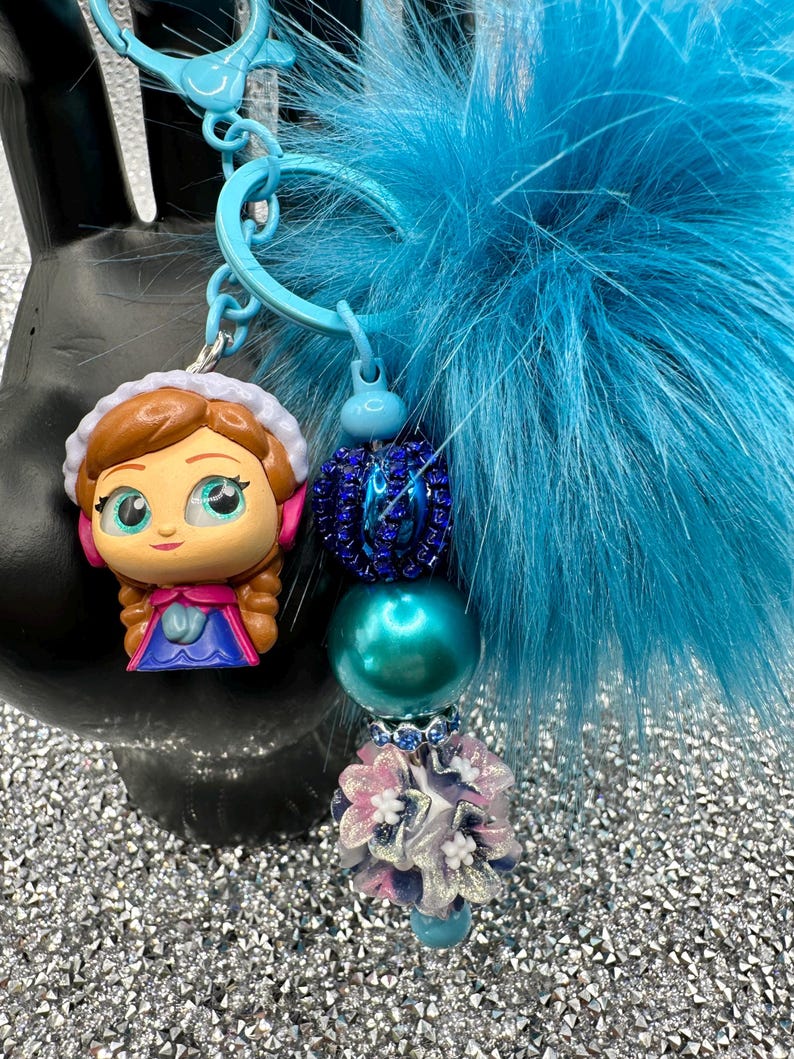 May include: A keychain featuring a cartoon character figurine with brown hair and a blue dress, a large bright blue faux fur pom-pom, and a strand of beads including a dark blue rhinestone bead, a teal pearl bead, and a floral-shaped bead.