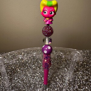 May include: A pink and yellow character with a white face and black eyes is on top of a pink and white glittery pen. The pen is in a clear plastic pen holder.