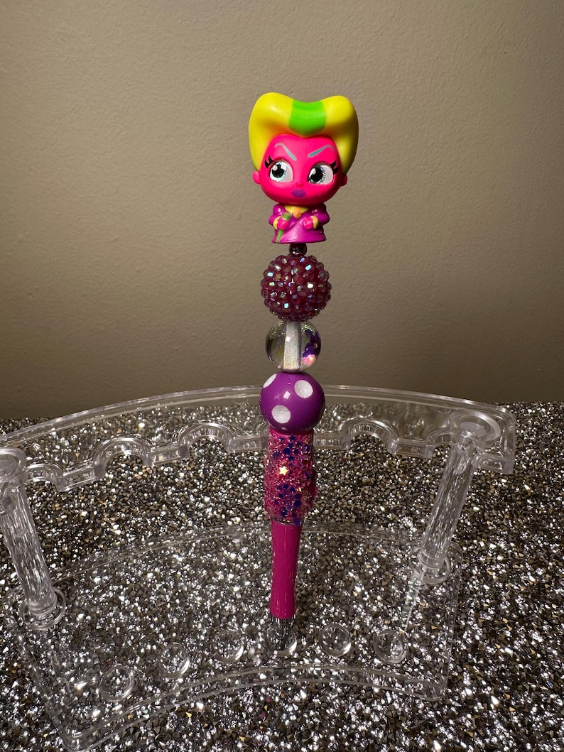 May include: A pink and yellow character with a white face and black eyes is on top of a pink and white glittery pen. The pen is in a clear plastic pen holder.