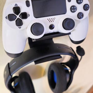 May include: A white and black gaming controller with a black headset. The controller has buttons and joysticks. The headset is black with blue accents. The controller and headset are mounted on a wall.
